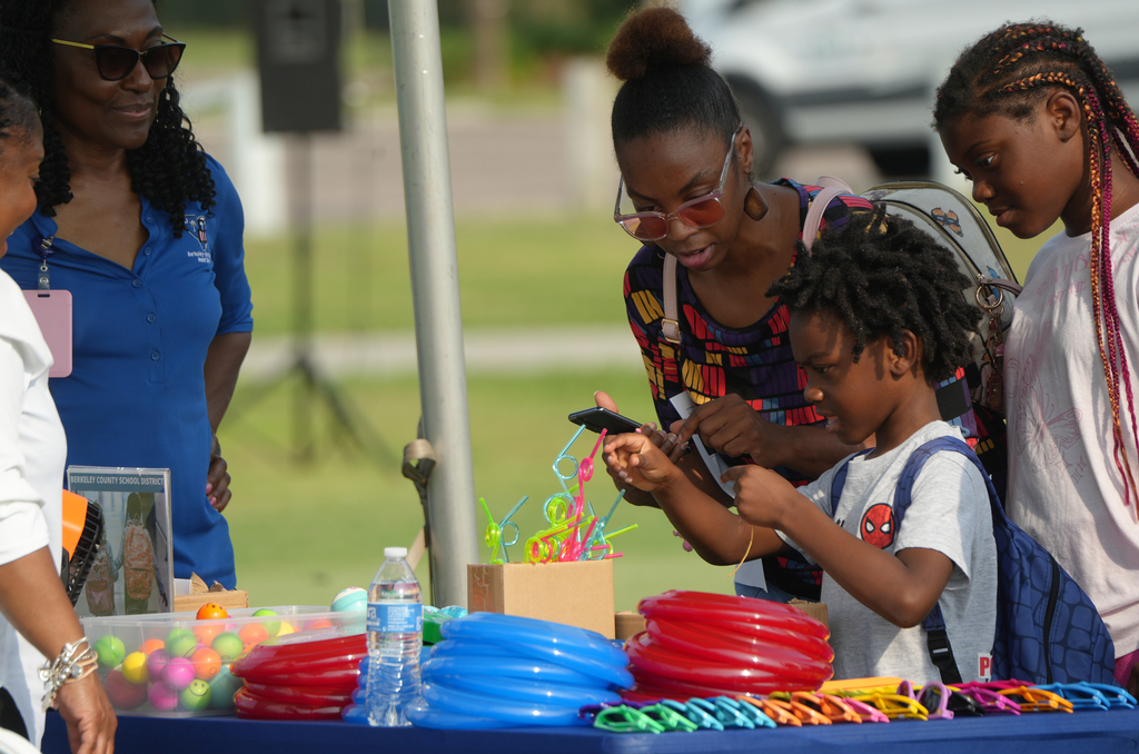 Four people gather at a table; one woman in glasses holds a phone, a child with a Spider-Man shirt uses it.