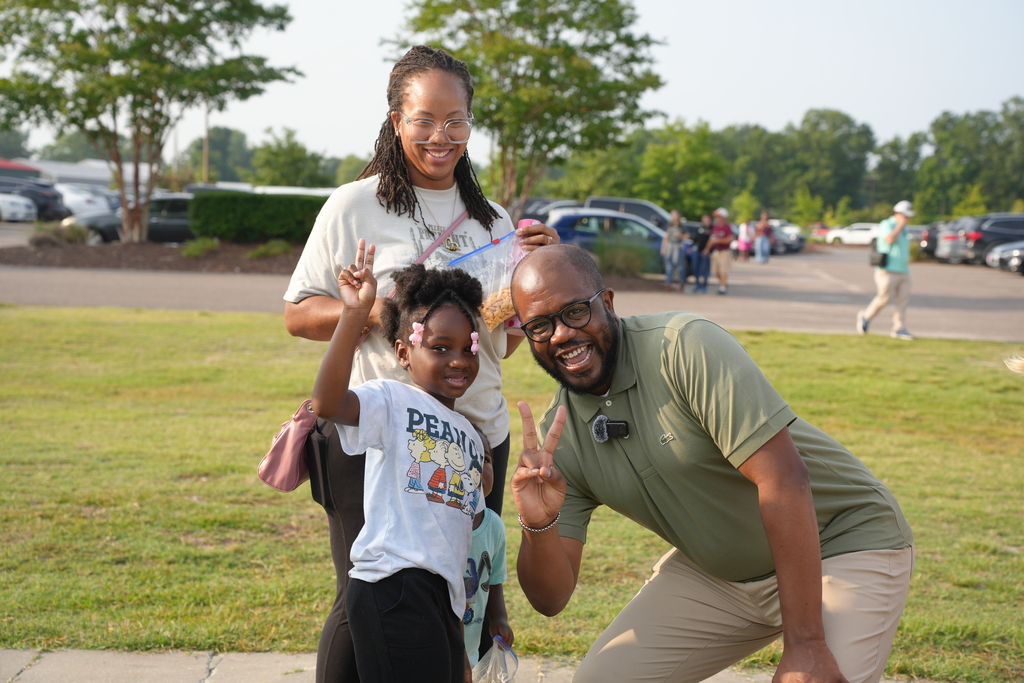 Three people on a grassy area with a parking lot in the background. Two adults smile at the camera.