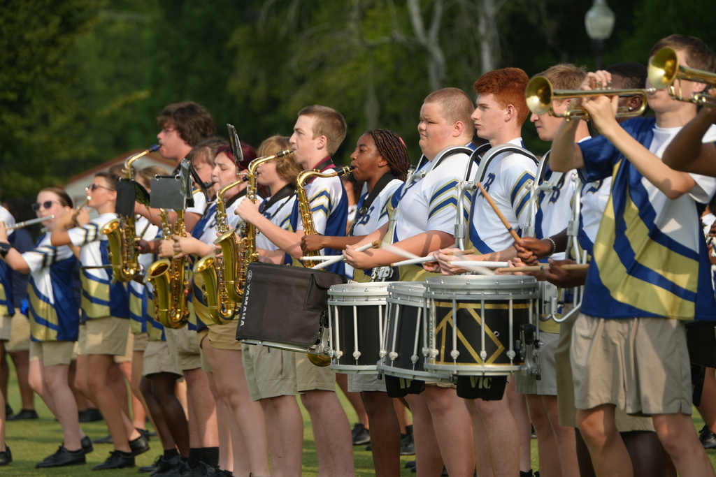 A group of students in blue and white uniforms plays trumpets, saxophones, and drums on a field.