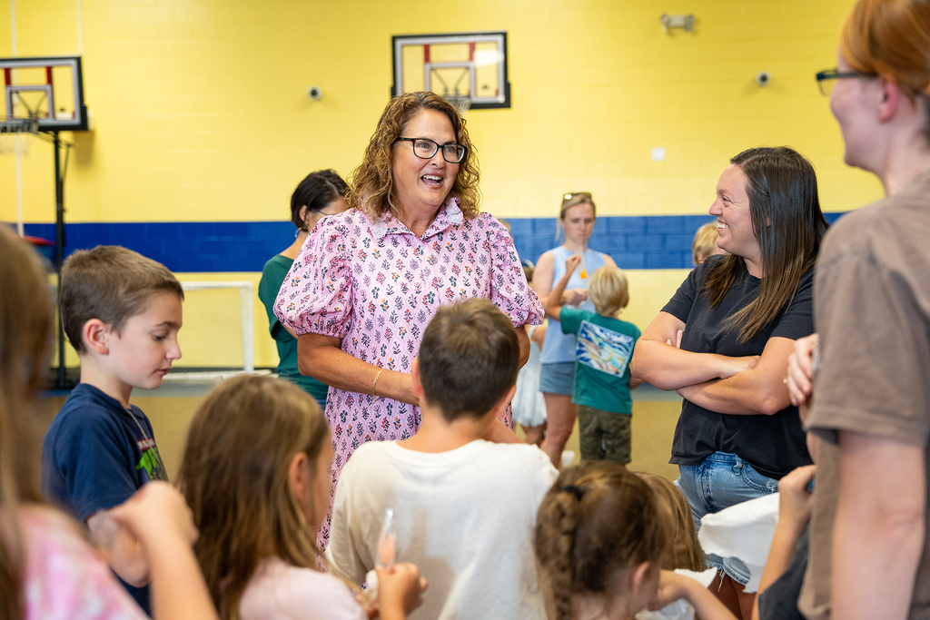 A group of children and adults gather indoors. A woman in a pink dress speaks to them.