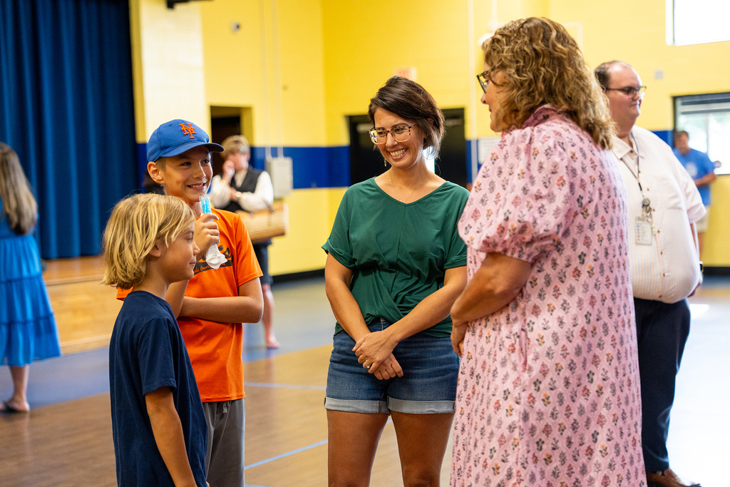 A group of people stands in a gym, with a young boy holding a microphone. A woman in a floral dress speaks to another woman in green.