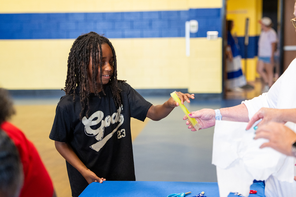 A young girl with curly hair smiles while holding a yellow object, standing at a blue table.
