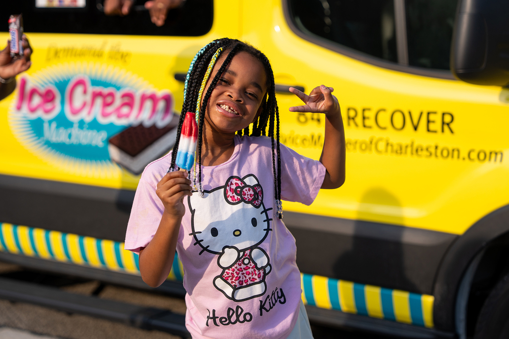 Girl with braids holding an ice cream popsicle in front of a yellow ice cream truck.