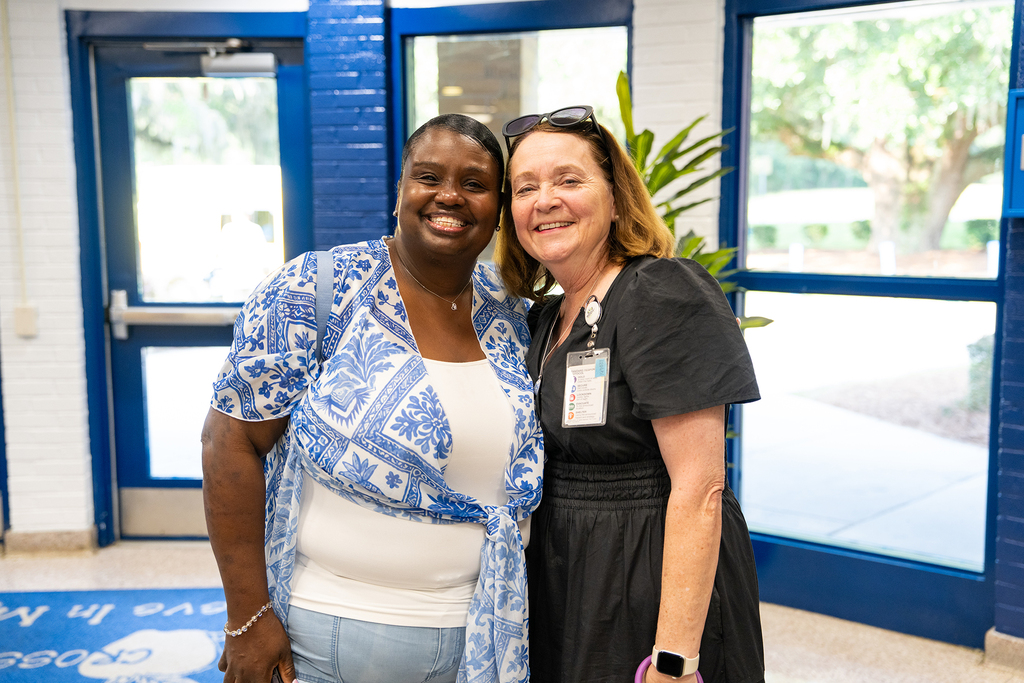 Two women smiling at camera, one in white and blue, the other in black, in a hallway with blue doors.