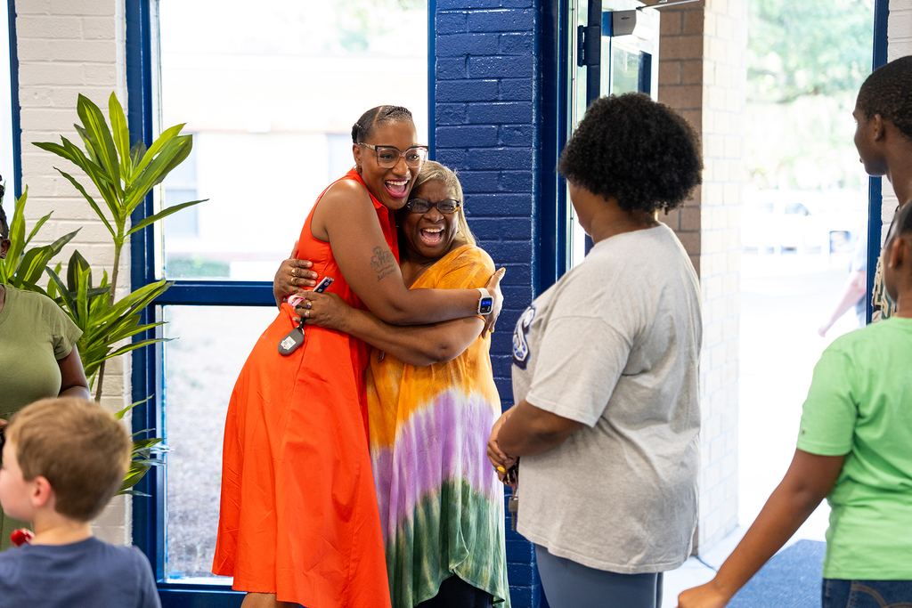 Adults and children gather in a room with large windows. Two women in vibrant dresses hug.