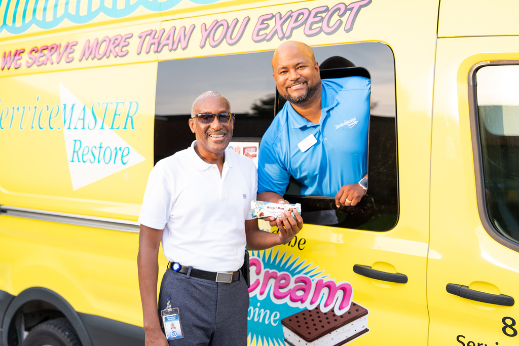 Two men are next to a yellow ice cream truck. One is standing, holding a container, and the other is inside.