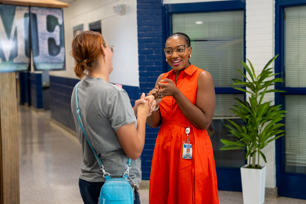 Two women, one in gray, the other in orange, face each other in a hallway. Blue bag, blue ID, and a potted plant are present.