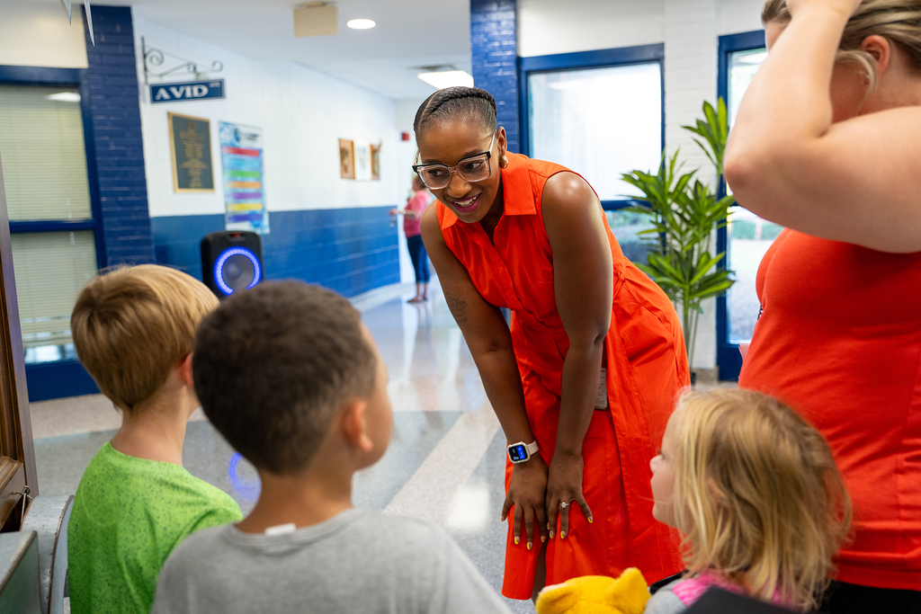 A woman in an orange dress stands with children in a hallway; a speaker is behind them.