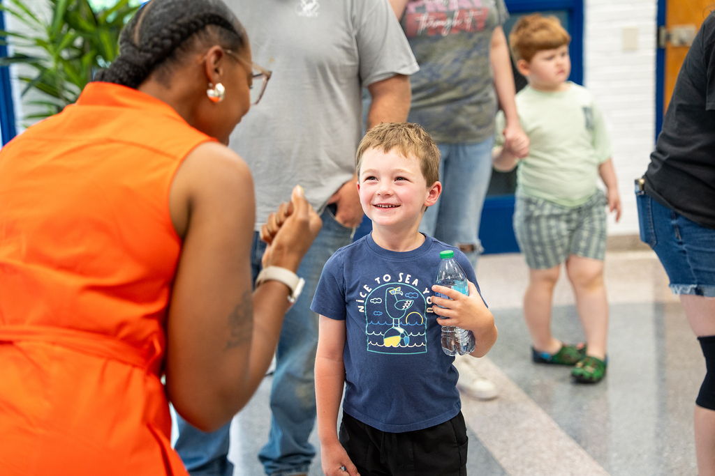 A young boy with a smiling face holds a bottle and stands next to a woman in an orange dress.