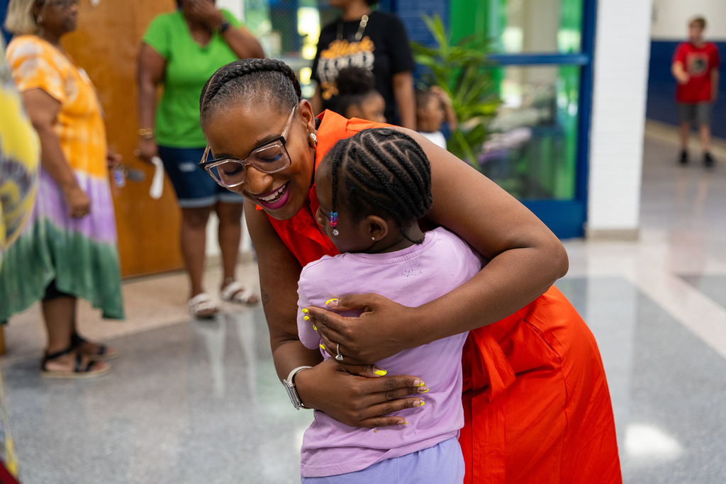 A smiling woman in a red dress holds a young girl in a purple shirt, both sharing a joyful hug.