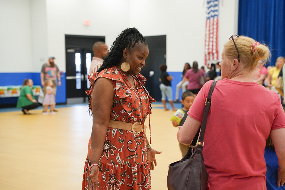 Two women converse, one in a patterned dress with a belt and the other in a pink shirt, in a gymnasium.