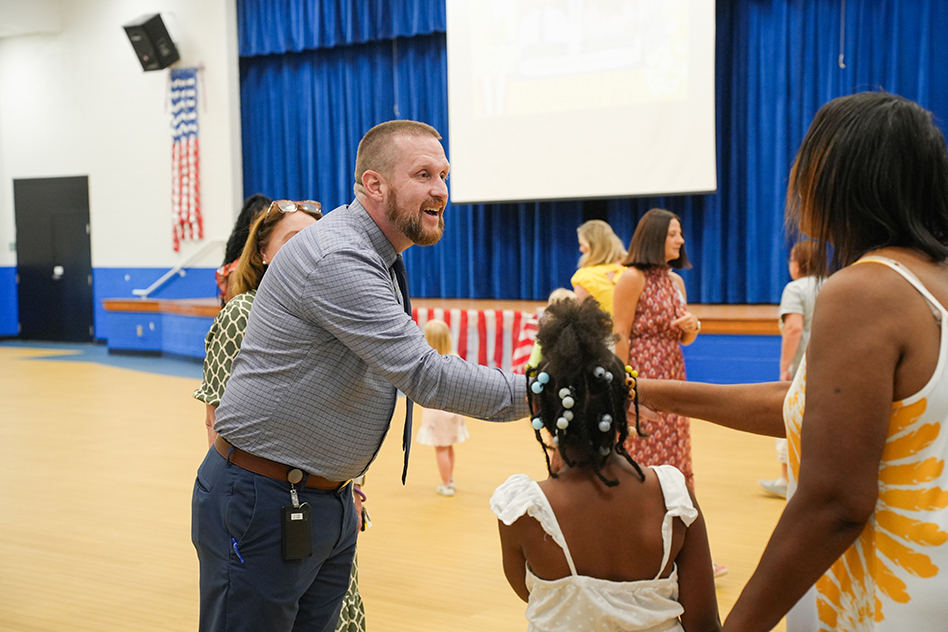 People in a room with a wooden floor and blue curtains, a man in a suit helps a child.