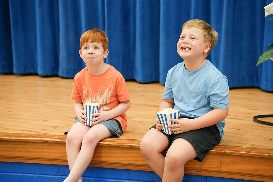 Two boys in orange and blue shirts, holding popcorn cups, sitting on a stage with a blue curtain in the background.