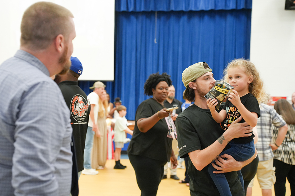 A group of people inside a gym, some holding children. A man carries a child on his shoulder, while others engage in conversation.