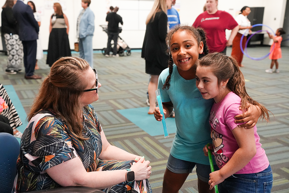 A woman and two girls stand together; the girls have hula hoops. A room full of people is in the background.