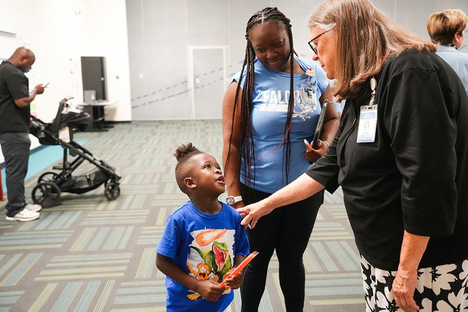 A woman and child look upwards as an older woman greets them in a room. A stroller and a man are in the background.