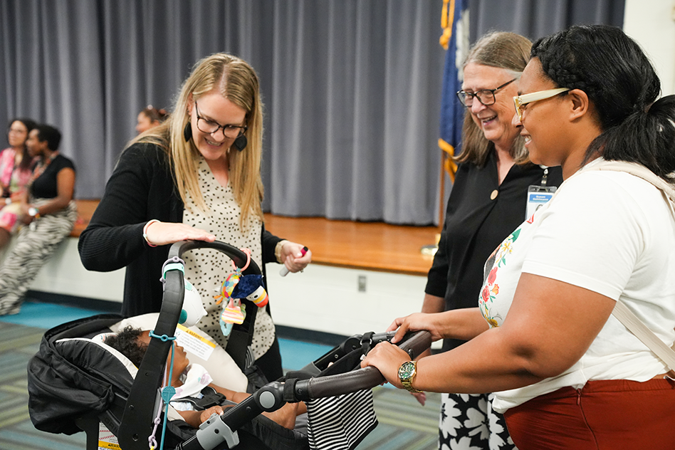 Three women interact with a baby in a stroller. One woman holds the stroller while another woman touches it.