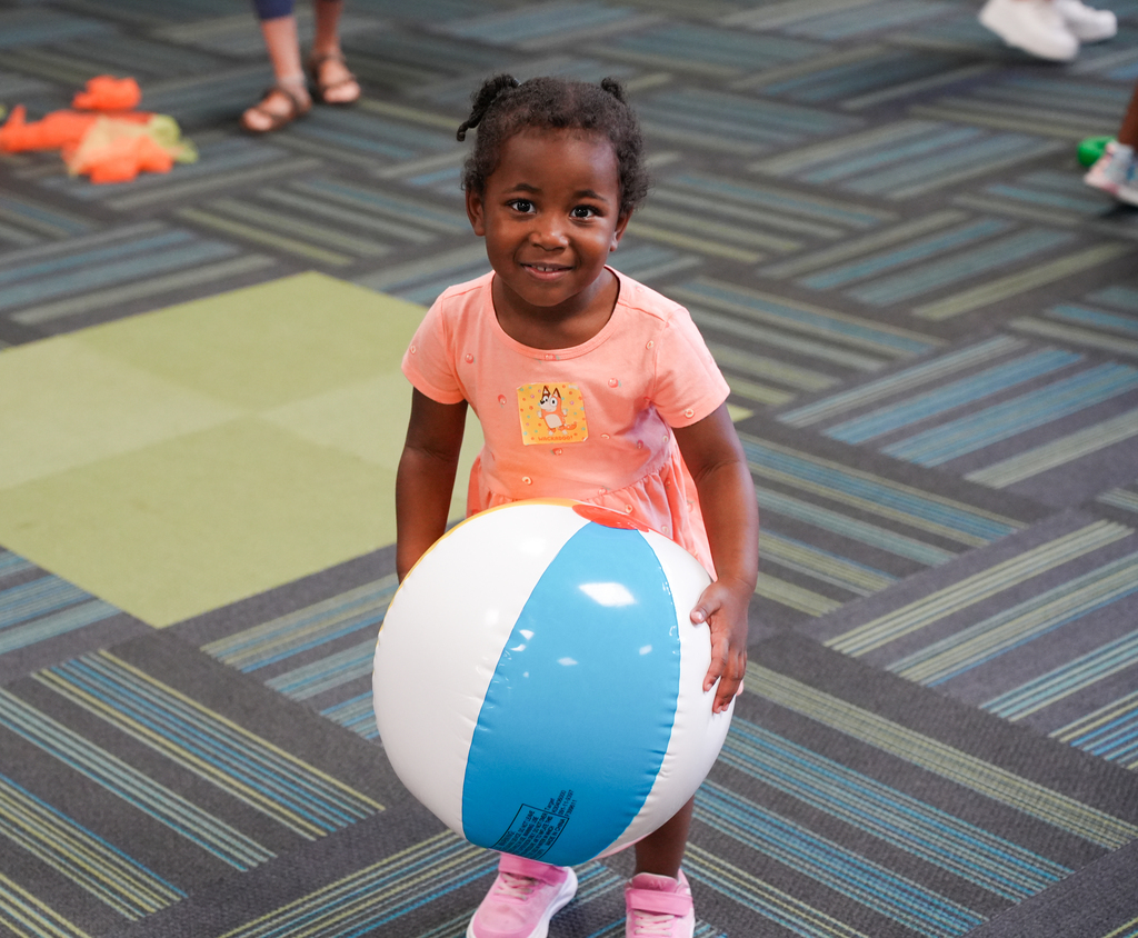 A young child, wearing a pink shirt, holding a large beach ball. In the background, a patterned floor and other toys.