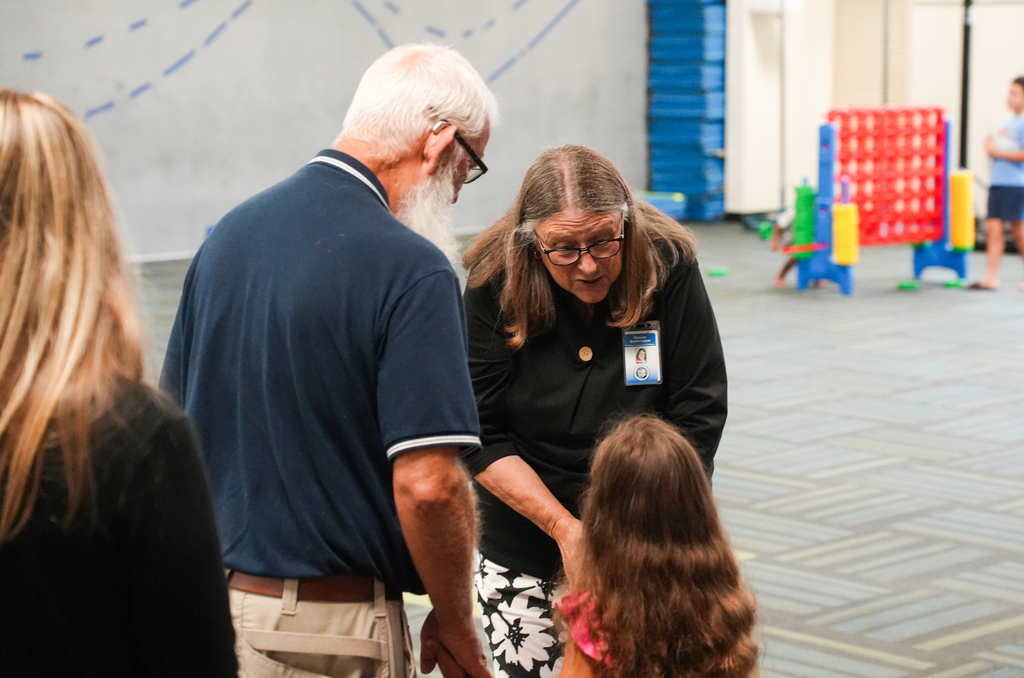 An older man and woman assist a young girl in a brightly colored room with colorful toys in the background.