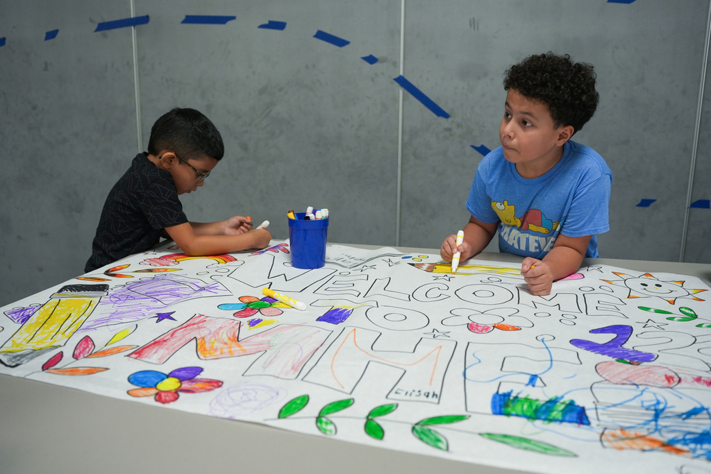 Two children sit at a table, drawing on a large sheet of paper. One wears glasses and a black shirt. The other wears a blue shirt.