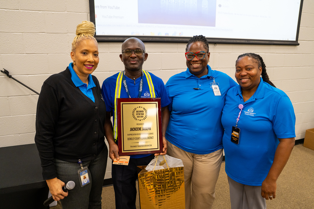 Four individuals stand in a room, one holding a framed award. They wear blue shirts, one wears a black top. A projector screen is in the background.