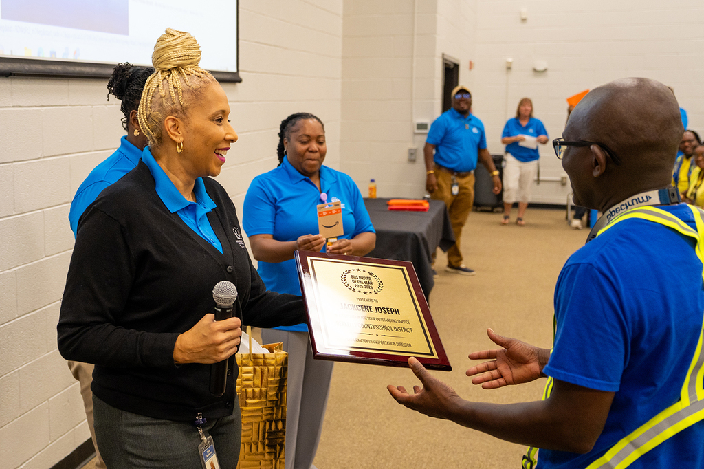 A woman hands a plaque to a man with a microphone in a room with others.
