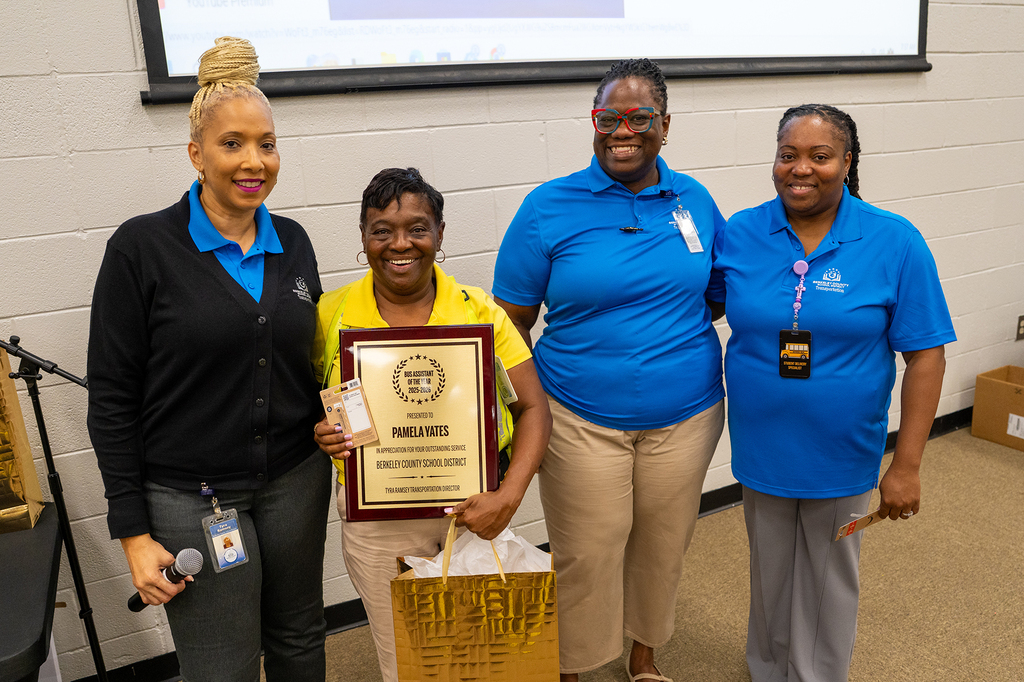 Four women in blue shirts stand in a room, one holding a plaque, another with a gift bag.