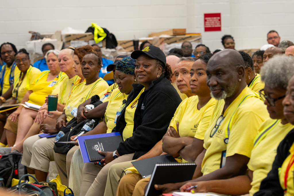 A crowd of people in yellow shirts sitting in rows, with some in dark clothing. Some have glasses, bags, and notebooks.