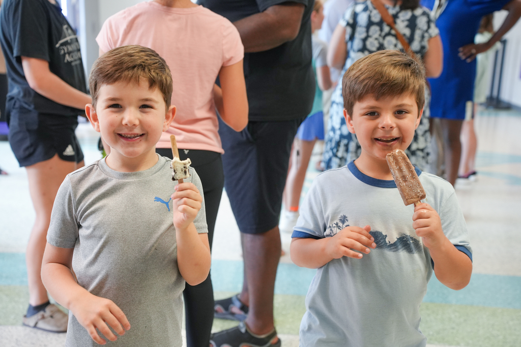 Two boys smile and show ice pops; one wears a gray shirt, the other a blue shirt. People stand behind them, one in a pink shirt and another in black shorts.