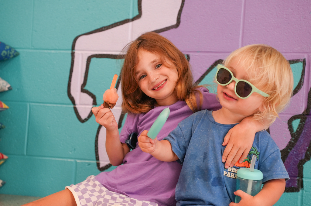 Two children hold ice pops, smiling and posing for a photo. One wears sunglasses and holds a cup.