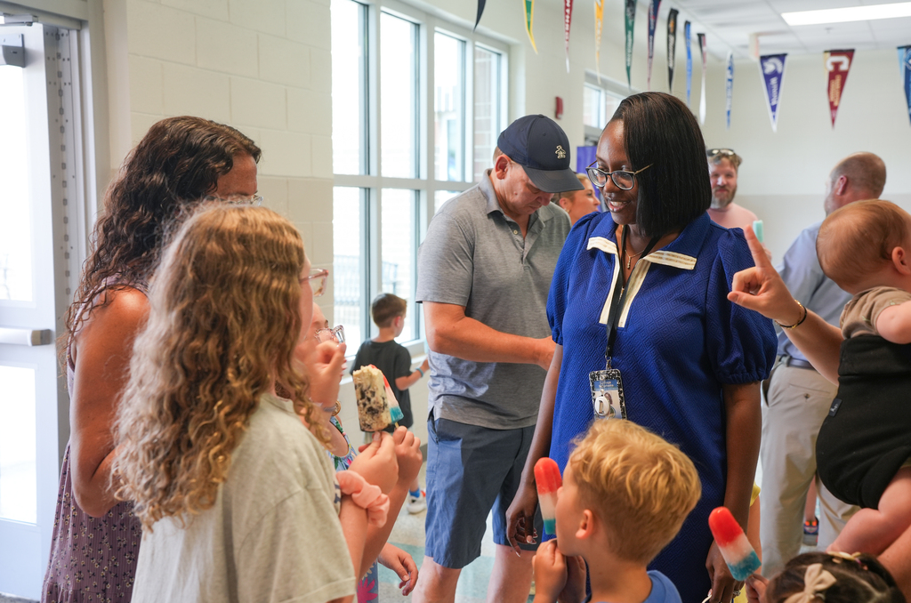 Several adults and children are gathered in a hallway. A woman in a blue shirt hands out ice pops.
