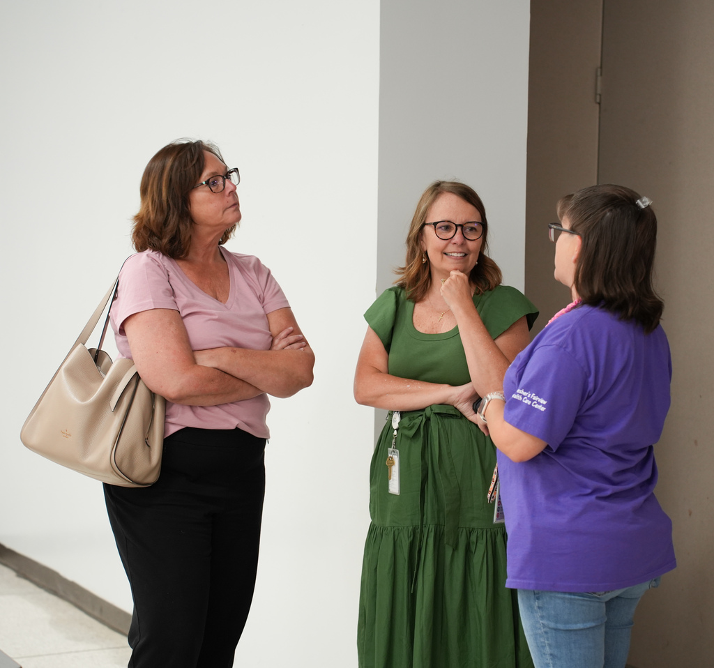 Three women stand in a hallway, engaged in conversation. One wears a pink shirt, another a green dress, and the third a purple shirt.