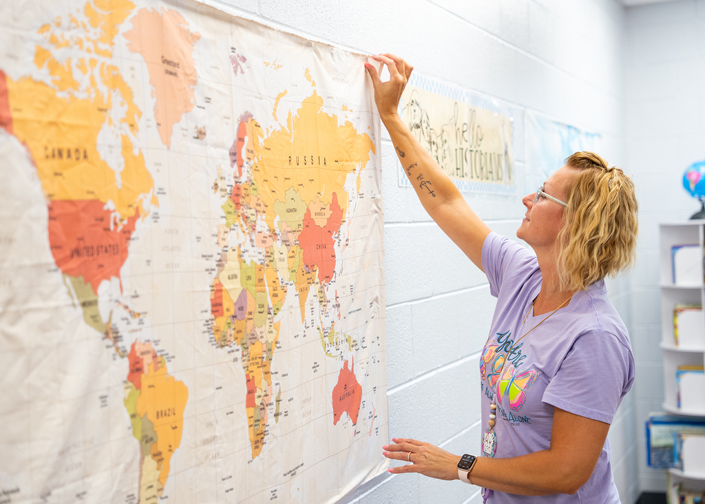 Woman holding up a world map with countries colored in different shades, standing in a classroom.