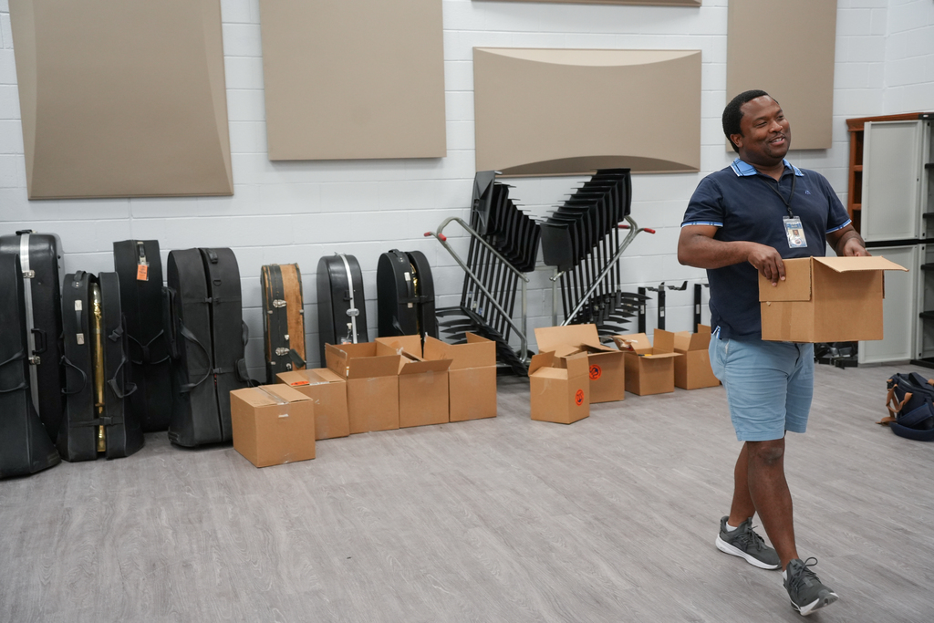 Man holding a box in a room with open boxes, black cases, and chairs on a gray floor.