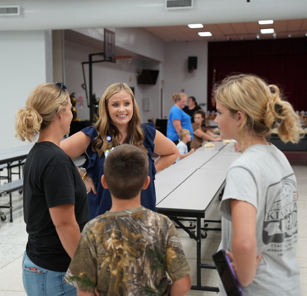 Four people, including two women and two children, stand in an indoor room. The women converse.