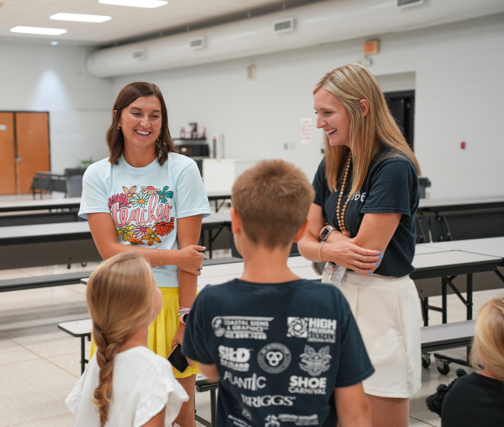 Two women and two children, one with a ponytail, talk in a room with a white ceiling and floor.
