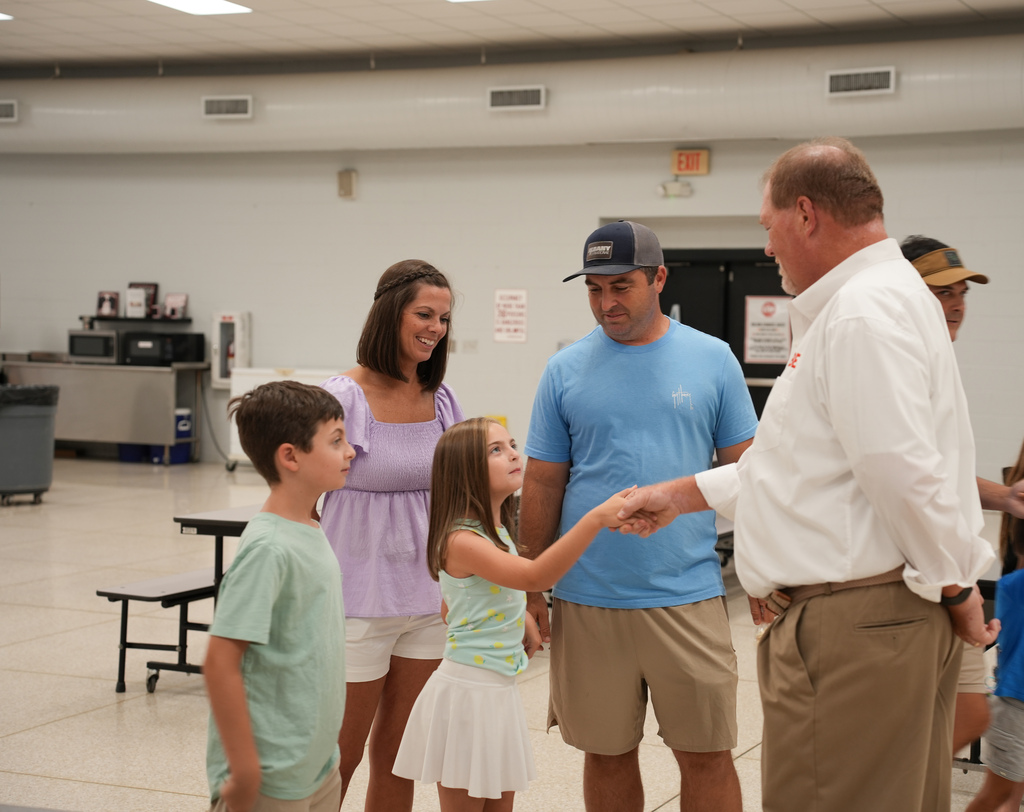A man shakes hands with a woman and child while two boys stand nearby. The room has white walls and lights.