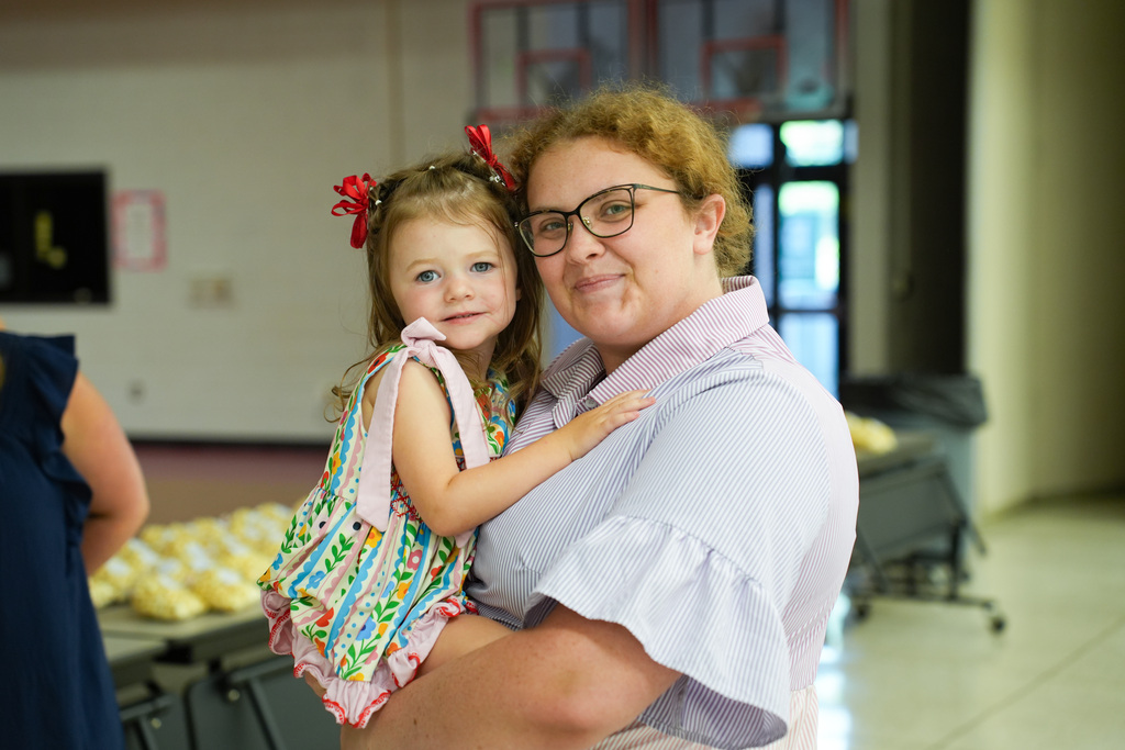 A woman holds a small child close. The child wears a dress with red bows, and the woman has glasses.