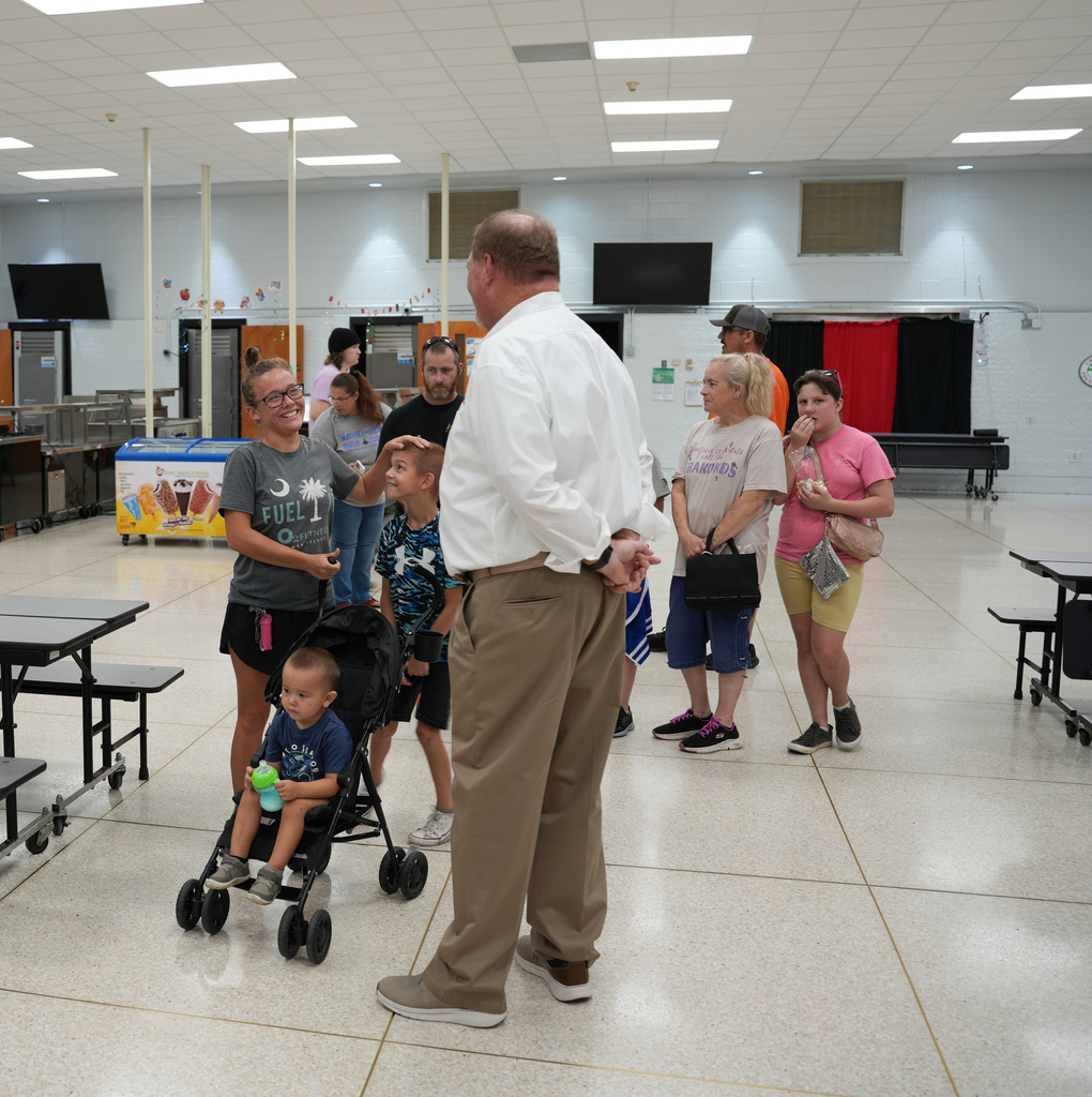 A room with a white ceiling, lights, tables, and people. A man speaks to others, some with children, in a cafeteria-style room.
