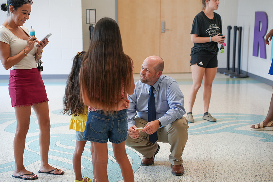 A man in a suit kneels while talking to a girl with long hair. Nearby, three women stand and look at their phones.