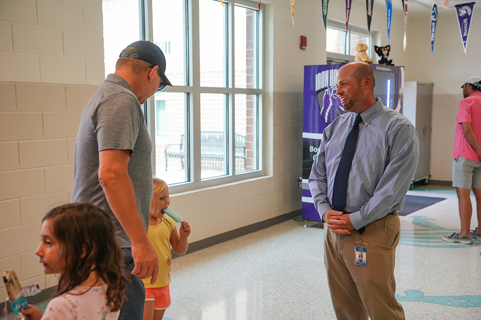 Two men converse in a hallway; one wears a cap and the other a tie. A girl looks at a book.