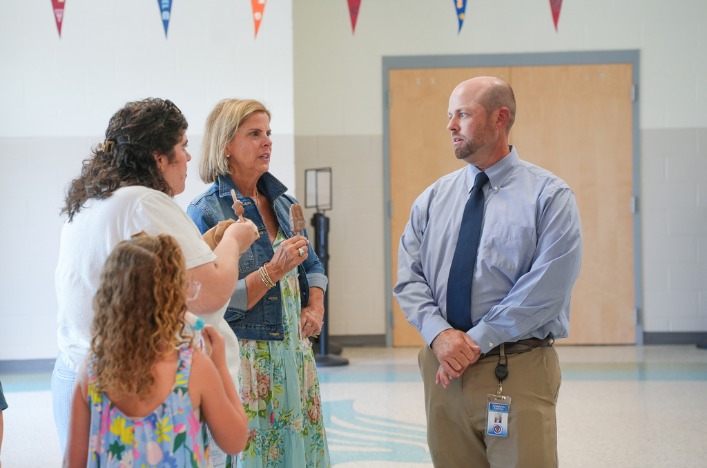 Four people in a hall; a woman holds a child's hand, another gestures, a man stands with folded hands, and a woman in a denim jacket talks.