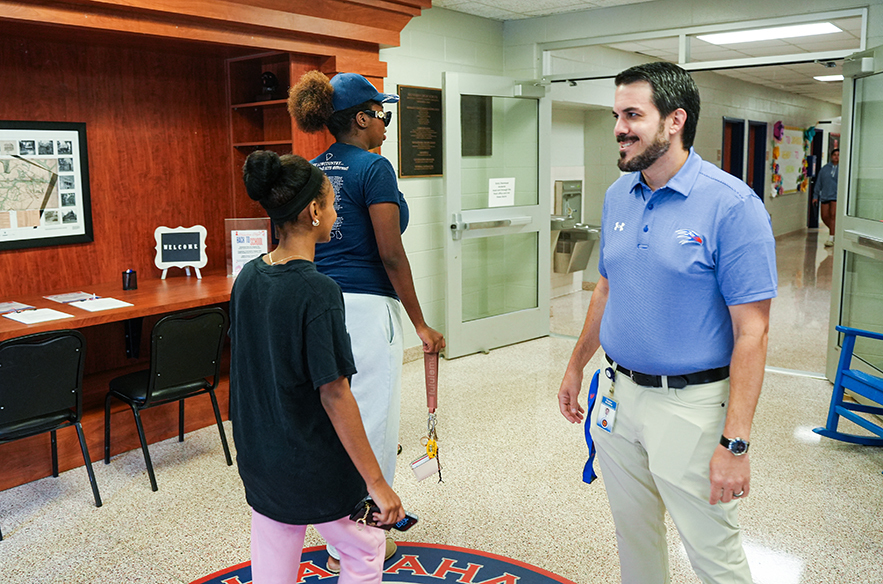Three people in a hallway; a man and a woman in blue shirts converse while a woman in black looks down.