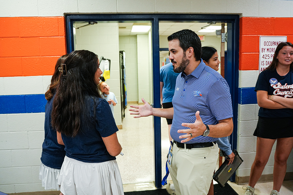 People gather in a hallway; a man speaks with others. He wears a blue shirt and pants. Some women wear white skirts, blue shirts, and sneakers.