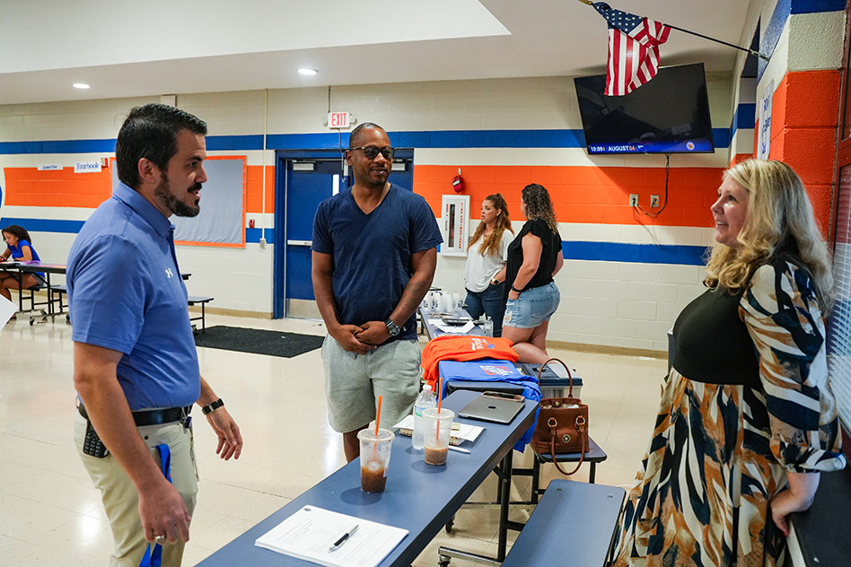 Four adults converse in a room with blue and orange walls. A flag hangs above a television.