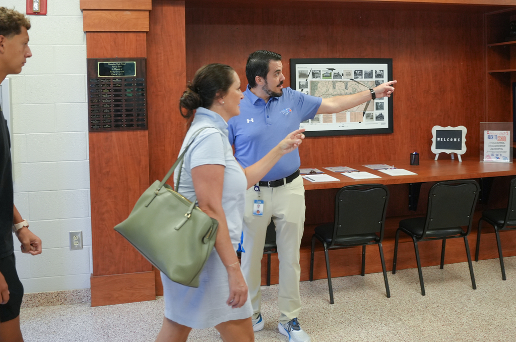 A man and a woman stand in a room, gesturing. A woman holds a green bag. Behind them, a man points to a framed picture on the wall.