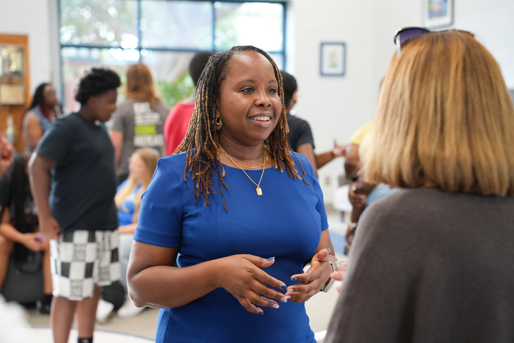 A woman in a blue shirt stands with hands gesturing as she talks to another woman in a gray shirt.