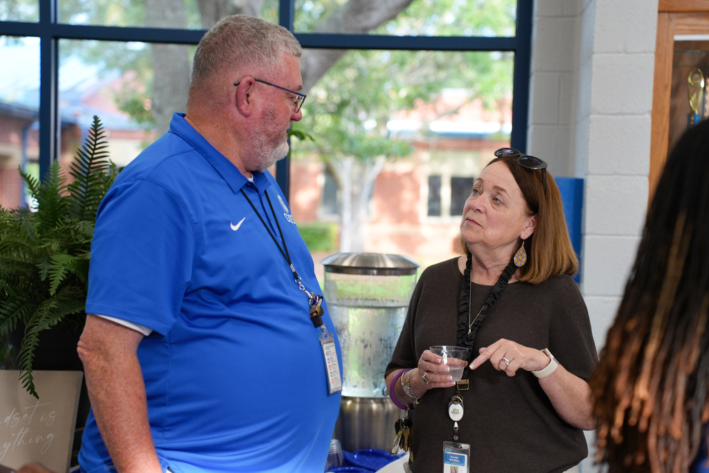 A man and a woman stand in a room, talking. The man wears glasses and a blue shirt, while the woman wears a brown shirt.
