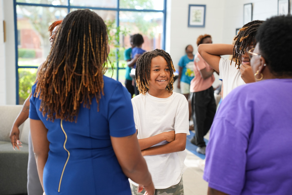 People inside a room; a child with braided hair smiles. Others wear glasses, and there are couches and windows.