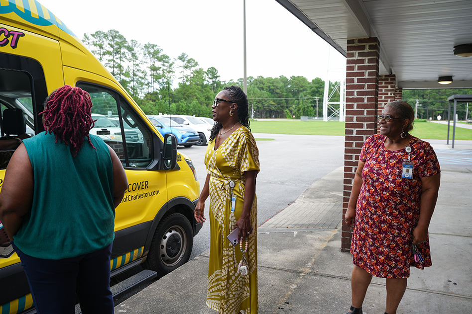 Three people stand by a yellow van. One woman in yellow and two others, one in green and one in red.
